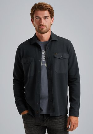 Dunkelgrau gemustertes Hemd mit texturierter Stoffoberfläche, zwei Brusttaschen und Kragen, über einem grauen T-Shirt getragen und mit dunklen Jeans kombiniert.