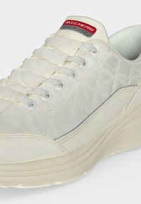 Baskets blanches Skechers avec motif matelassé, lacets blancs et semelle épaisse, affichant l'étiquette de la marque sur la languette.