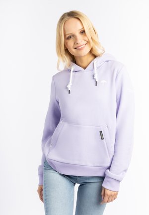 Jonge vrouw die glimlacht, draagt een lichtpaarse hoodie met trekkoorden en lichtblauwe spijkerbroek tegen een witte achtergrond.