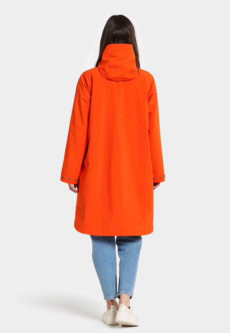 Orangen wasserfester Regenmantel mit Kapuze, ausgestattet mit langen Ärmeln, geradem Schnitt und Druckknopfdetails an den Bündchen. Getragen über blauen Jeans.