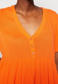 Helles oranges Kleid mit V-Ausschnitt, kurzen Ärmeln und einem gestuften Saum. Drei Knöpfe zieren die Vorderseite, aus leichtem strukturiertem Stoff gefertigt.