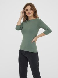 Ung kvinde med brunt hår iført en grøn tætsiddende bluse med trekvartærmer og sorte jeans, poserer med hånden på hoften og smiler.