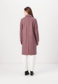 Lange geruite jas met een rood en blauw ruitpatroon, op maat gesneden en met een geplooide kraag. Gecombineerd met witte broeken en zwarte haklaarzen.