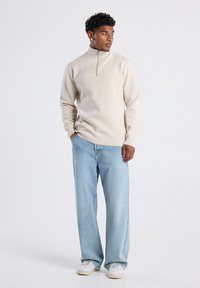 Beige quarter-zip fleece pullover, relaxed fit, with ribbed cuffs. Light blue loose-fitting jeans and white sneakers complete the outfit.