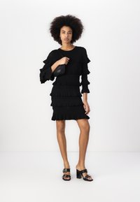 Svart kjole med lagdelte volanger, lange ermer og en tettsittende fasong. Modellen bærer en svart clutch og svarte hælede sandaler med spenne.