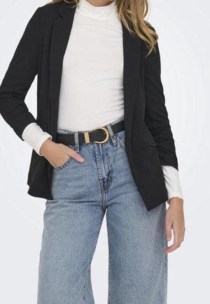 Femme portant un blazer noir sur un col roulé blanc, rentré dans un jean taille haute bleu clair avec une ceinture noire à boucle dorée.
