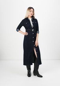 Femme portant une longue robe en denim foncé à boutons avec une ceinture, une chemise blanche en dessous et des bottes noires montantes, se tenant devant un fond blanc.