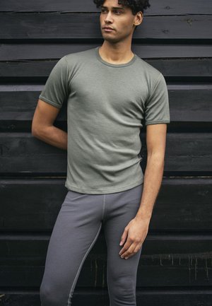 Olivenfarvet t-shirt med kortærmer lavet af blødt stof, sammen med tætsiddende grå leggings, mod en sort trækulisse.