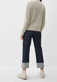 Hellbeiger Strickpullover mit figurbetontem Schnitt und strukturiertem Muster, kombiniert mit dunkelblauen Jeanshosen mit umgeschlagenen Bündchen und cremefarbenen Turnschuhen.