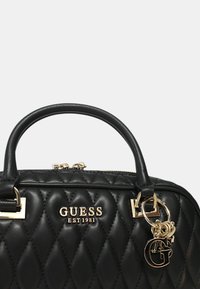 Sort quiltet håndtaske med guldfarvet hardware, præget "GUESS EST1981" logo og en dekorativ charm. Har dobbelt håndtag og lynlås lukning.