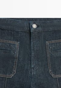 Dunkle Jeans aus Denim mit einem sichtbaren Metallknopf an der Taille, zwei Gesäßtaschen mit oranger Naht und einer glatten, strukturierten Oberfläche.