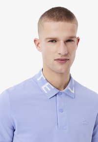 Lichtpaarse polo-shirt van textuurstof, met een witte kraag voorzien van logo en een klein geborduurd logo op de borst.