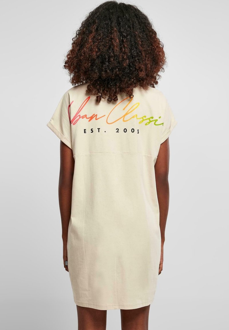 Personne aux cheveux bouclés porte une robe beige clair à manches courtes avec un texte coloré "Urban Classic EST. 2005" dans le dos.