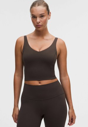 Kvinde iført en tætsiddende mørkebrun ærmeløs crop top og matchende højtaljede leggings, stående mod en ensfarvet lys baggrund.