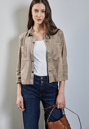 Beige Wildlederjacke mit zwei Fronttaschen, über einem weißen eng anliegenden Top und hoch taillierten dunkelblauen Jeans getragen. Braune Handtasche in der rechten Hand gehalten.