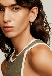Mujer que lleva una camiseta sin mangas de color oliva con ribete blanco, collar de plata y pequeños pendientes de aro con un detalle de gema transparente.