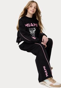 Conjunto de sudadera y pantalones negros con la palabra "MIAMI" en letras rosas. Cuenta con piping lateral y estampado gráfico. El material parece suave y cómodo.
