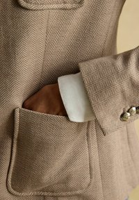 Main dans la poche d'un blazer beige en chevrons avec un poignet de chemise blanc et trois boutons métalliques sur la manche.