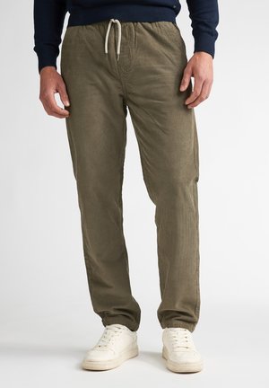Man draagt olijfgroene corduroy joggingbroek met trekkoord in de taille en witte sneakers, staand tegen een effen achtergrond.