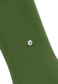 Zbliżenie na zieloną skarpetę z małą, okrągłą, srebrną metką z czarnym tekstem, podkreślające fakturę i kolor materiału.