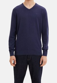 Mann trägt einen schlichten dunkelblauen V-Ausschnitt-Pullover mit langen Ärmeln und dunkle Jeans, steht vor einem weißen Hintergrund.