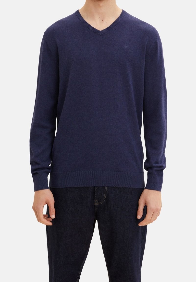 Mann trägt einen schlichten dunkelblauen V-Ausschnitt-Pullover mit langen Ärmeln und dunkle Jeans, steht vor einem weißen Hintergrund.