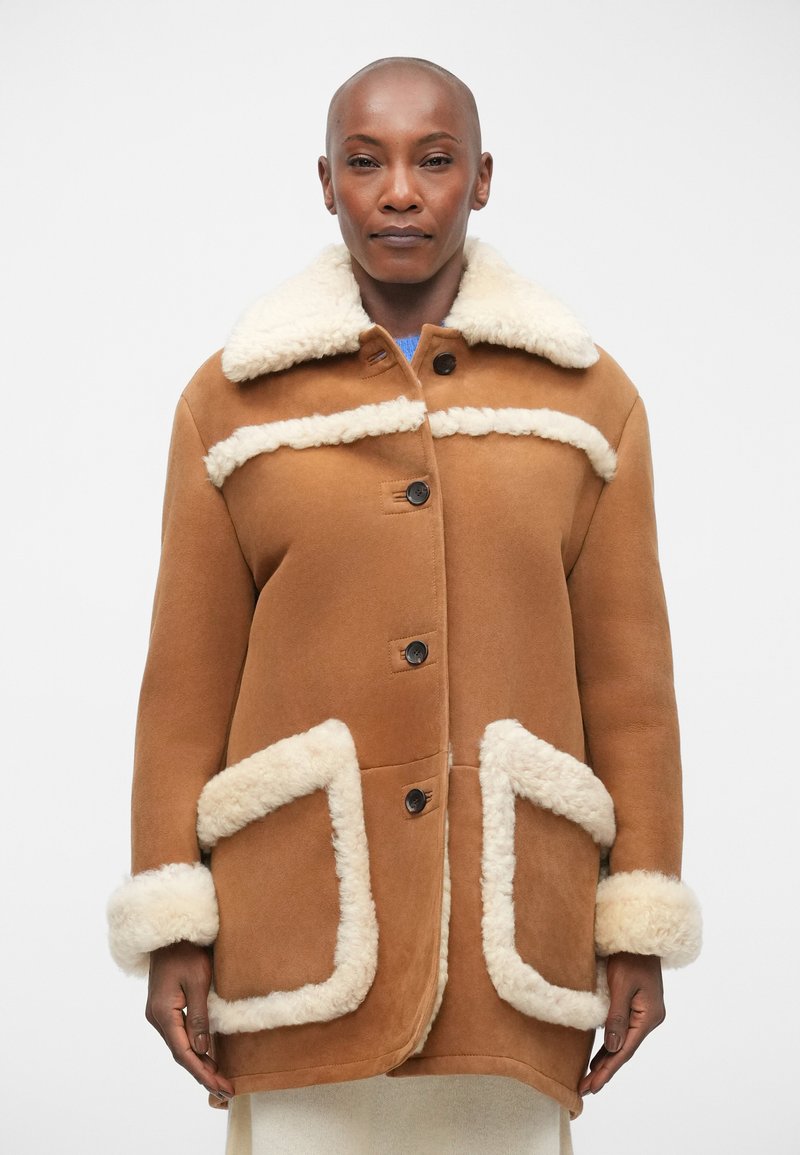Cappotto in camoscio marrone con bordi in pelliccia sintetica crema, chiusura a bottoni e due tasche a patta. Presenta un colletto e una vestibilità comoda.