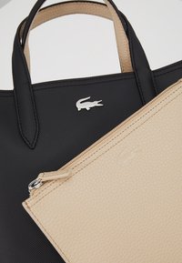 Sac cabas noir texturé avec poignées et logo crocodile argenté, pochette zippée beige texturée chevauchant le sac cabas.