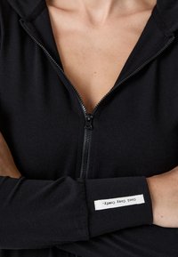 Persona vestida con una prenda negra de cierre con una etiqueta blanca en la manga que dice "Cool Cosy Comfy", con los brazos cruzados sobre el pecho.