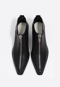 Botas de cuero negro hasta el tobillo con punta afilada y una cremallera plateada prominente en la parte frontal, con una textura suave y un diseño minimalista.