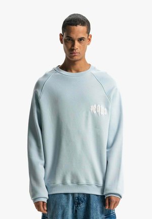 Lys blå sweatshirts med rund hals og raglanærmer. Har et subtilt hvidt logo på brystet og en tekstureret stoffinish.