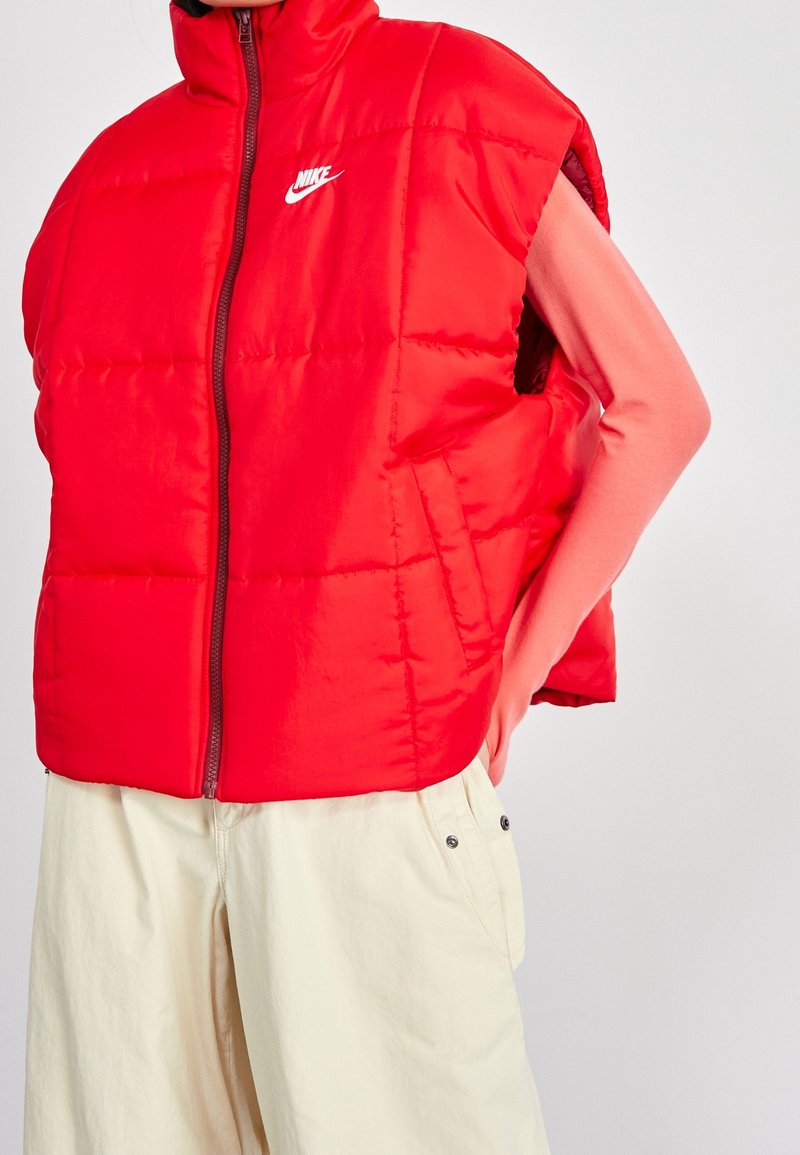 Röd vadderad väst med hög krage och framsida dragkedja, med en signatur vit logotyp. Tillsammans med ljusa, löst sittande byxor.