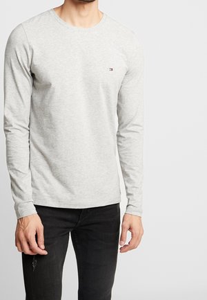 Mężczyzna ubrany w jasnoszary, długorękawowy sweter z małym logo na piersi oraz czarne dżinsy z niewielkim rozdarciem na lewym udzie.