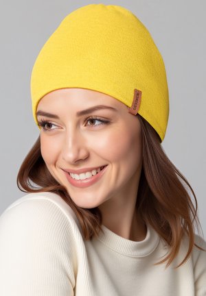 Gelbe Strickmütze mit einer strukturierten Oberfläche, die an der Seite ein kleines braunes Lederetikett aufweist. Getragen mit einem hellen Pullover.