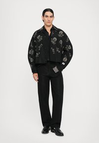 Schwarze Cropped-Jacke mit floralem Bestickung, kombiniert mit schwarzen strukturierten Hosen. Die Jacke hat einen Kragen, Knöpfe an der Vorderseite und ein Etikett.