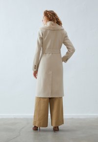 Beige wool coat with a faux fur collar, belted waist, and buttoned cuffs. Features a straight silhouette and center-back vent.