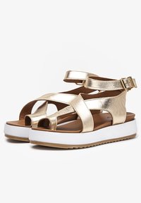 Złote metaliczne sandały z krzyżującymi się paskami, otwartym noskiem i białą platformą z teksturowanym beżowym spodem. Regulowane zapięcie na klamrę.