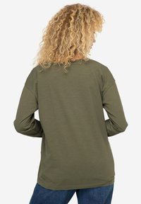 Oliwkowa koszula z długim rękawem o luźnym kroju, miękkiej fakturze i opadających szwach ramion. Pokazana z tyłu, w połączeniu z niebieskimi dżinsami.