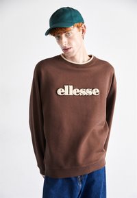 Sudadera marrón con un logo bordado "ellesse" en color crema, con un cuello de canalé. Combinada con unos jeans de mezclilla azules y una gorra color teal.