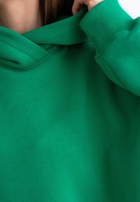 Felpa verde con una texture morbida, presenta un cappuccio a doppio strato e dettagli a coste sui polsini. La vista ravvicinata mette in risalto il colore e il materiale.