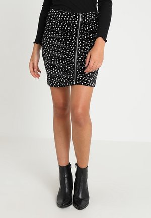 Nainen, joka on pukeutunut mustavalkoiseen pilkulliseen minihameeseen, jossa on etuvetoketju, mustaan pitkähihaiseen toppiin ja mustiin nilkkureihin, seisoo.