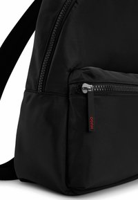 Coin d'un sac à dos noir montrant une poche zippée avant avec une languette de tirette noire portant l'inscription "HUGO" en lettres rouges et une bretelle rembourrée.