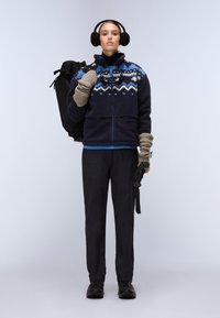 Giacca in pile con motivo blu e bianco, pantaloni neri, stivali neri e guanti. Porta uno zaino grande nero. Presenti i copriorecchi.