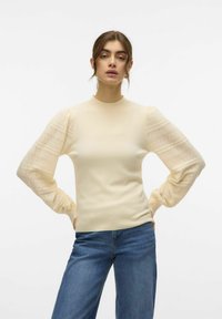 Lichtbeige getailleerde trui met pofmouwen van kant, voorzien van een textuurpatroon, ronde halslijn en geribde manchetten. Gecombineerd met blauwe jeans.