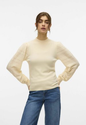 Lichtbeige getailleerde trui met pofmouwen van kant, voorzien van een textuurpatroon, ronde halslijn en geribde manchetten. Gecombineerd met blauwe jeans.