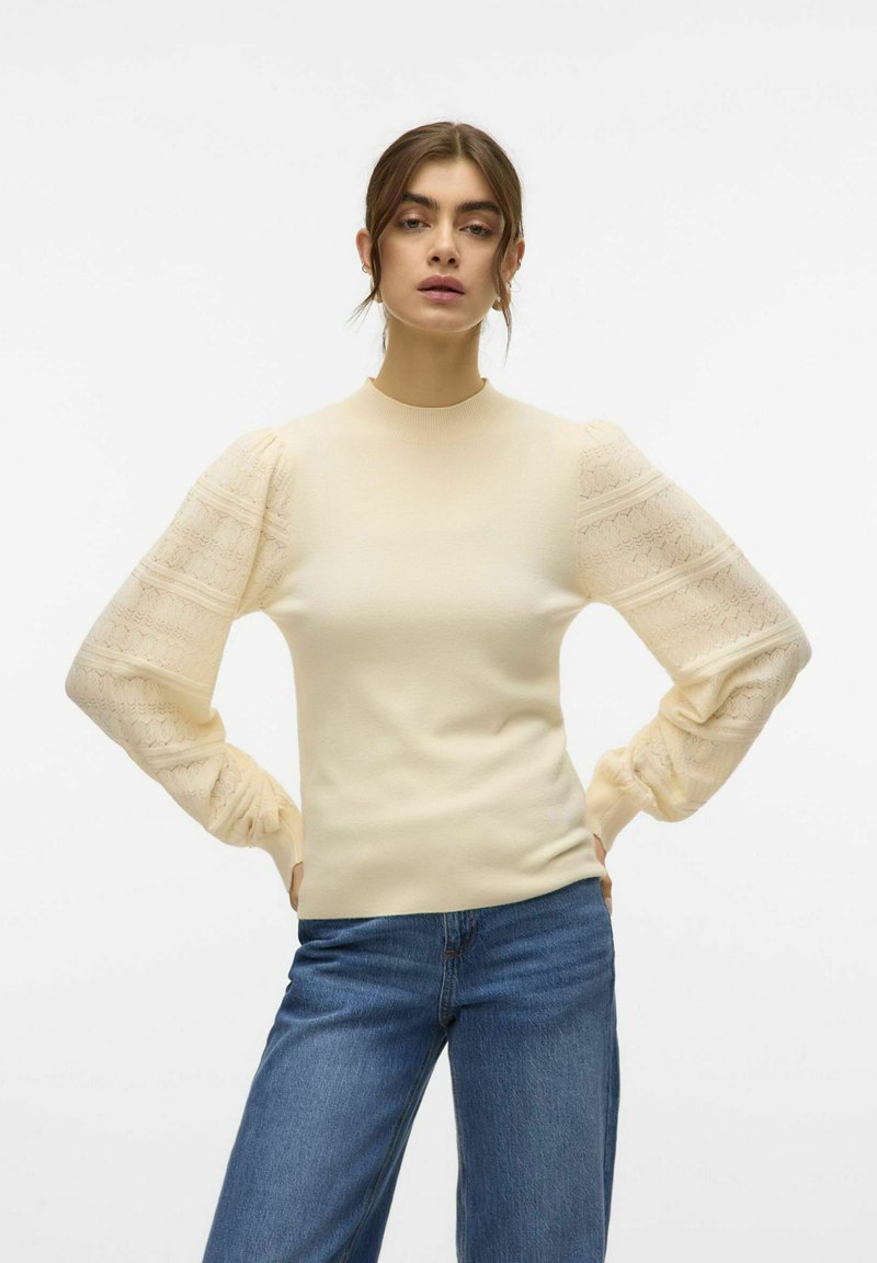 Lichtbeige getailleerde trui met pofmouwen van kant, voorzien van een textuurpatroon, ronde halslijn en geribde manchetten. Gecombineerd met blauwe jeans.