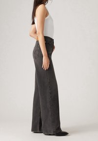 Džíny z černého denimu s širokými nohavicemi a vysokým pasem, s kapsami na zadní straně a mírným třepením na lemu, v kombinaci s bílým tílkem.