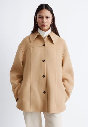 Frau trägt einen beigen, geknöpften Wollmantel mit großem Kragen, weißen Rollkragenpullover, weiße Hose und goldene Creolen, die Hände in den Manteltaschen.