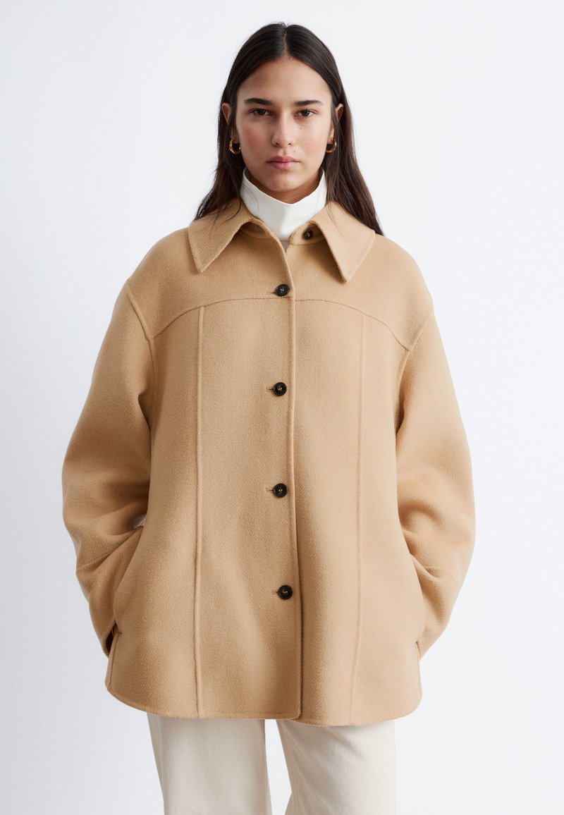 Kvinde iført en beige uldfrakke med knapper og stor krave, hvid rullekrave, hvide bukser og gyldne øreringe i hoops, hænderne i frakkens lommer.