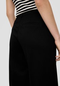 Schwarze weit geschnittene Hose aus strukturiertem Stoff mit seitlichen Taschen und einem glatten Bund. Vertikal gestreiftes Oberteil ist teilweise sichtbar.
